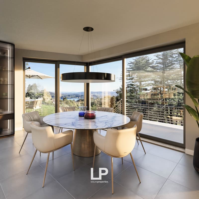 Spacious dining area with a round marble-top dining table and beige upholstered chairs. Large windows provide a scenic view of the outdoor landscape. A modern pendant light fixture hangs above the dining table.