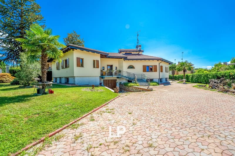 Two-story house, beige exterior, brown roof, large driveway, large yard, trees, garden, staircase, door in the garage