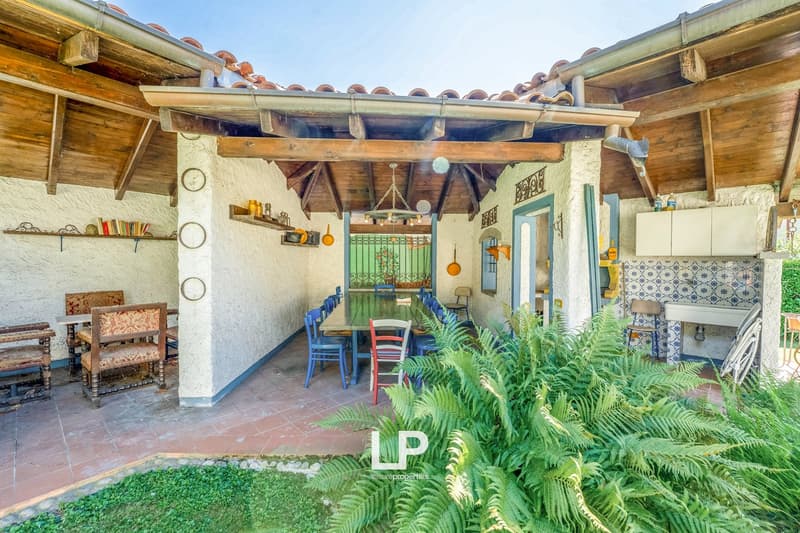 Outdoor dining area with a table and chairs, wooden beams on the ceiling, a green awning, tiled floor, and decorative wall shelves with items like bottles and a green curtain.