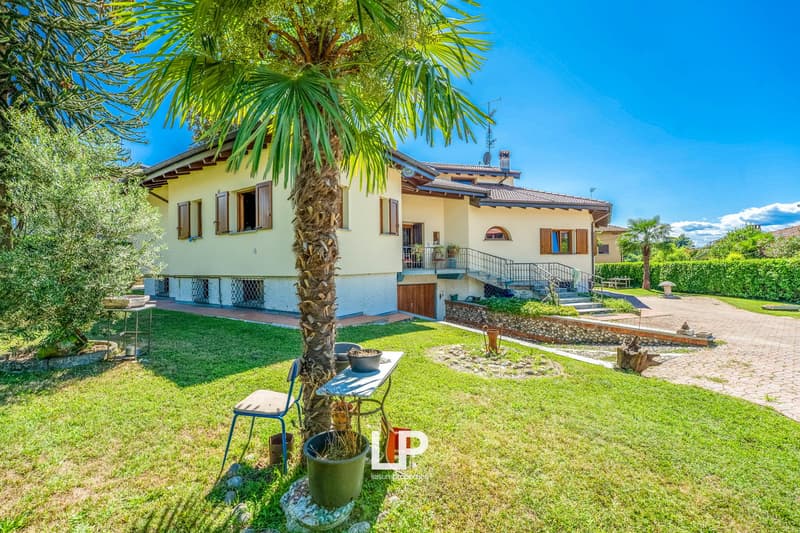 Two story house, yellow facade, brown tiled roof, multiple windows, wooden entrance door, stairs to entrance, well-maintained garden, tall palm tree, small table, garden chairs, green bushes, clear view