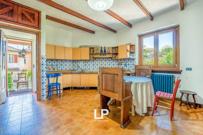 Open kitchen with dining area, wooden cabinets, blue tiled wall, ceramic floor, table with white tablecloth, wooden benches and chairs, open door to balcony, radiator.