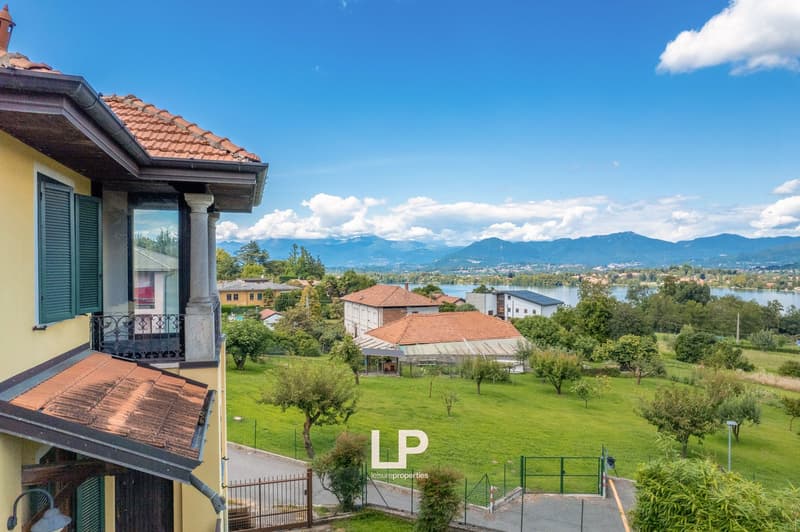 Terraced house, red roof, yellow facade, balcony with railings, fenced garden, water body in background, mountains in distance
