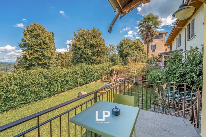 A balcony with a table and chairs, surrounded by a metal railing, overlooking a garden with greenery and bushes, and a clear blue sky in the background.