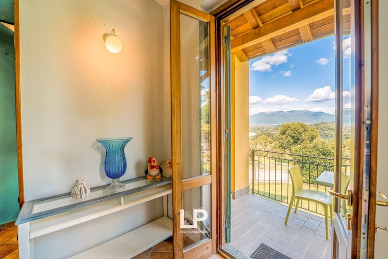 Interior room with white walls, wooden door, and a large glass door leading to a balcony with railings. A shelf on the left has decorative items and a blue vase. On the balcony, there is a chair, a small table, and a view of the surrounding greenery and m