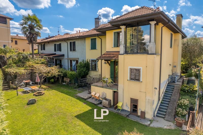 two-story house with yellow walls, tiled roof, glass window with balcony, garden with green lawn, patio with chairs and table, decorative rocks and fire pit