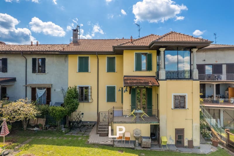 Two story house, yellow exterior, green shutters, balcony, staircase leading to terrace, lush garden, sunny weather, distant buildings in the background