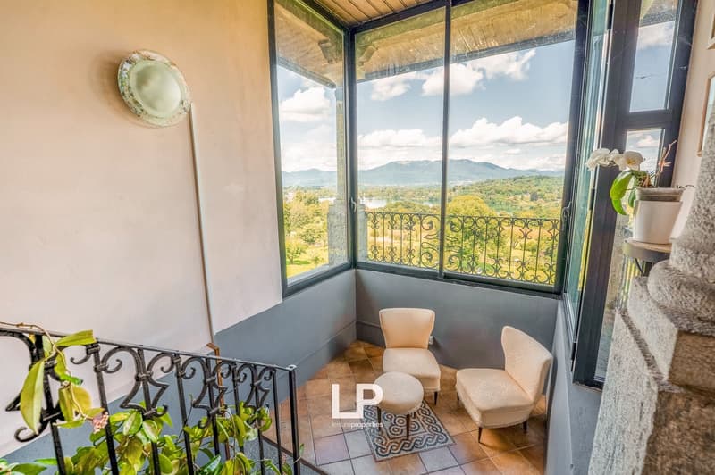 Balcony with large windows, iron railing, beige wall, tile floor, chairs, table, greenery, plant pot, distant view of hills and lake.