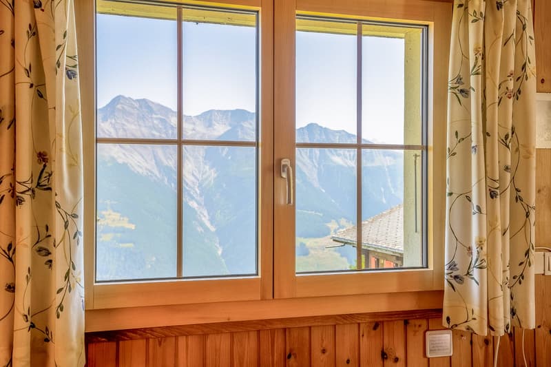 Large window, wooden frames, curtains with floral pattern, view of mountains and houses