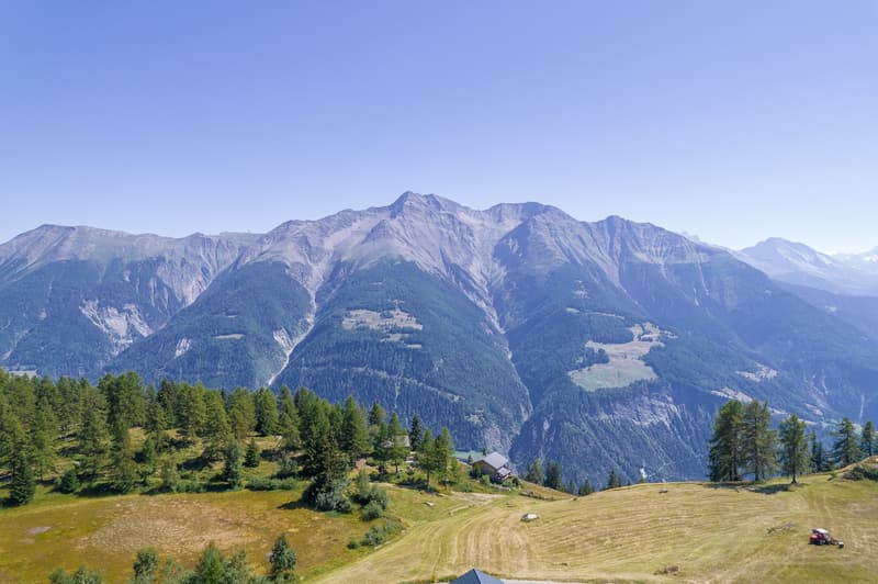 Wide aerial shot of mountains with houses, fields, and a tractor in the foreground with trees in the distance under a clear blue sky.