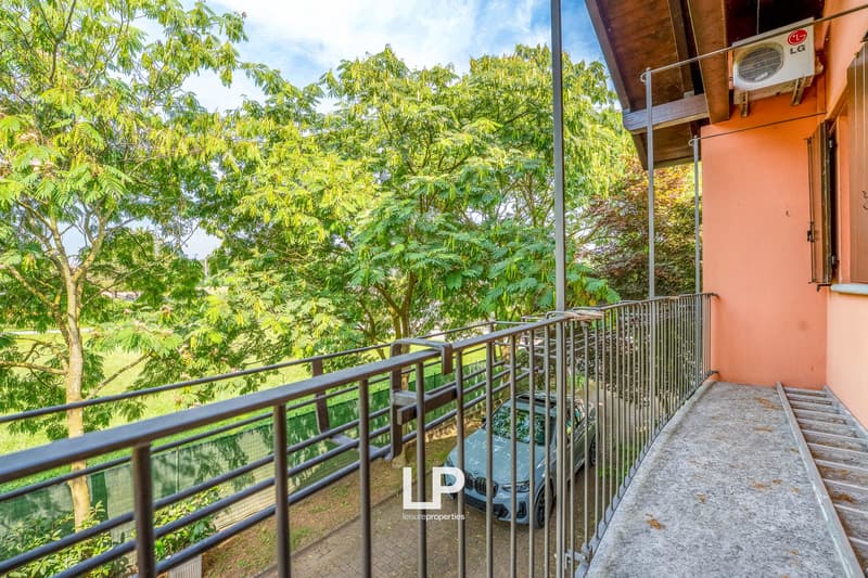 Car parked in front of a balcony, black railings, tree with green leaves, grass field, air conditioning unit