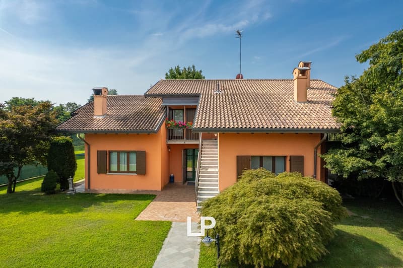 Two-story house, orange exterior, terracotta roof, stairs leading to the entrance, front yard with green lawn, garden with bushes and flowers, clear sky background.
