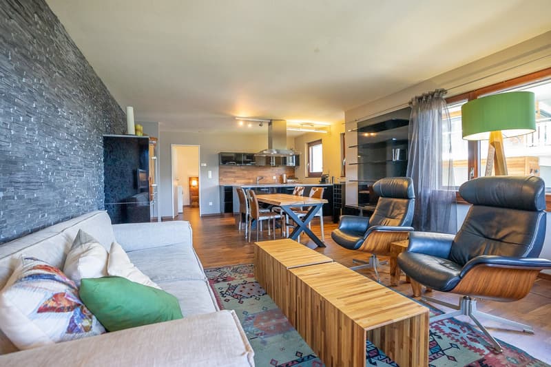 open plan living and dining area, gray couch with cushions, wooden coffee table, blue rug, two armchairs, lamp, dining table with chairs, kitchen island with bar stools, gray wall with stone texture