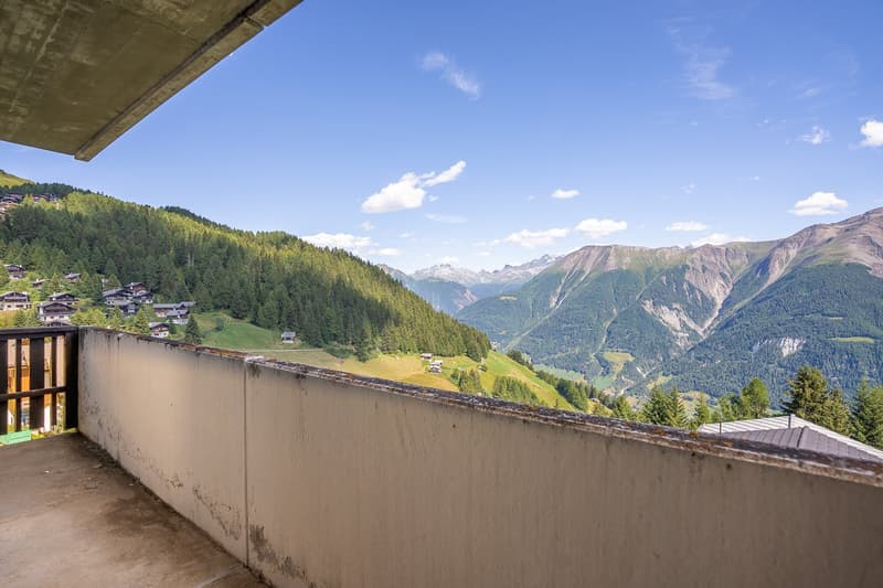 A balcony with wooden railing, a wall and a view of a mountain range with villages, trees, and a blue sky with clouds.