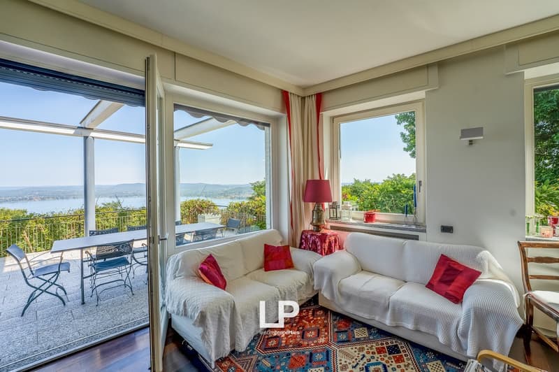 A room with large windows, an open door to a balcony with an outdoor table and chairs, a couch covered with a blanket, a coffee table, and a lamp.