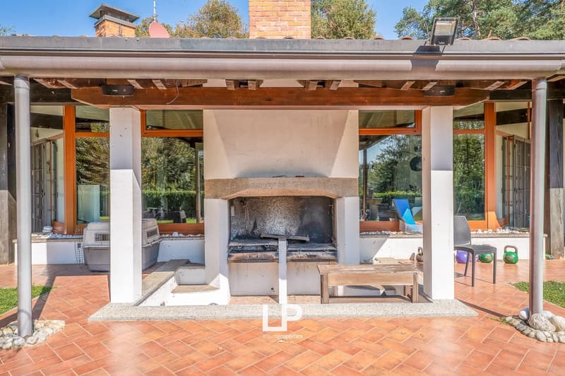 Covered patio with brick and stone elements, outdoor fireplace, large glass windows, wooden bench, outdoor chair, open pet kennel, tiles