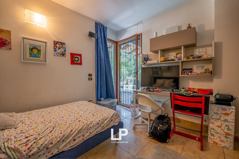 small bedroom with bed, desk, chair, bag, monitor, shelf with various objects, blue curtain