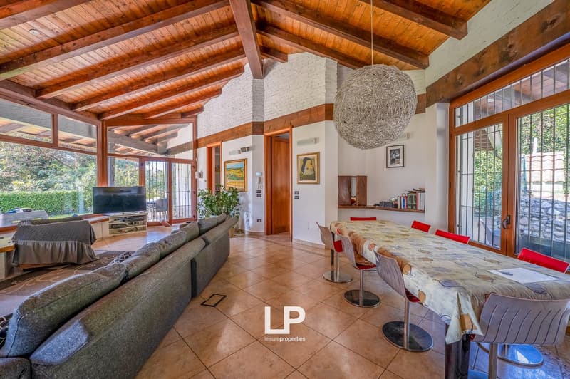 large living room, wooden ceiling, multiple large windows, long dining table, chairs, gray sofa, television, large chandelier, wooden furniture