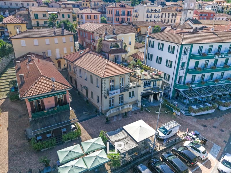 An aerial view of a town, featuring various buildings with terracotta roofs and balconies, nestled among trees and greenery. Streetlamps, cars parked, and an open-air cafe are visible in the foreground. The setting suggests a small town with historical ar