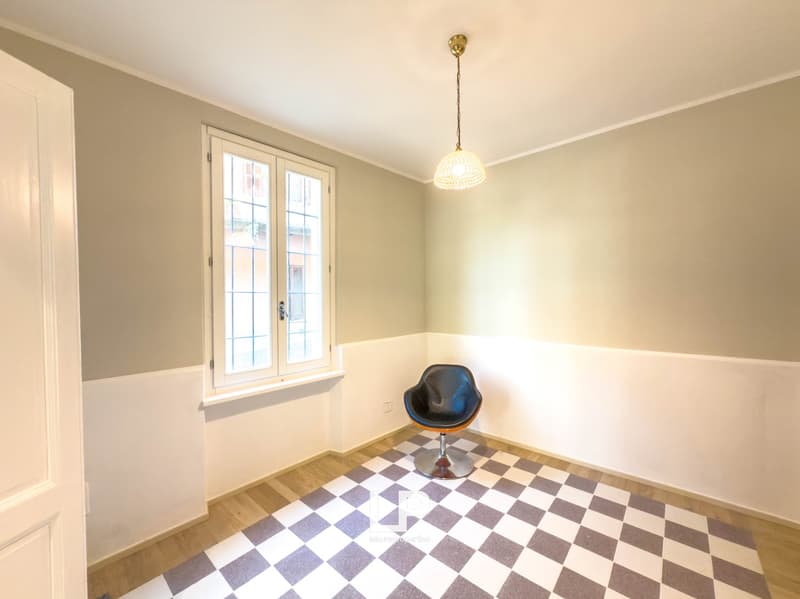 Empty room with wooden floor, white and grey checkered flooring, gray wall, white door, small black chair, light fixture