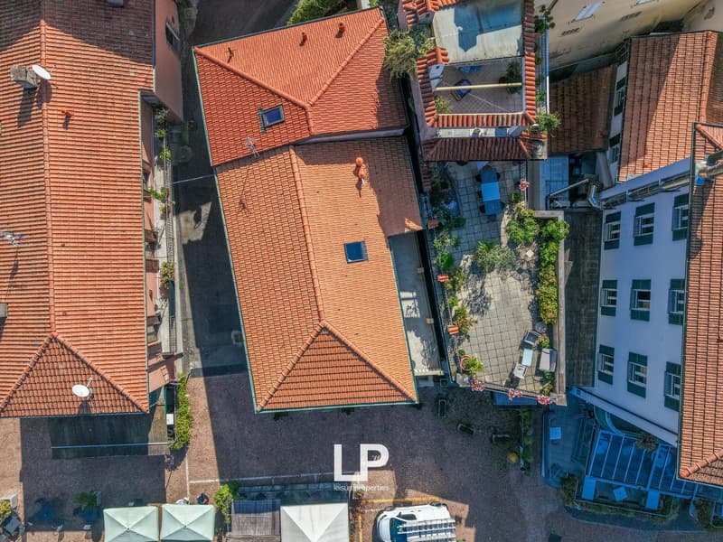 Buildings with red tiled roofs, some featuring skylights, situated in a compact urban setting, surrounded by small courtyards and a paved street, with a white van parked in the foreground.