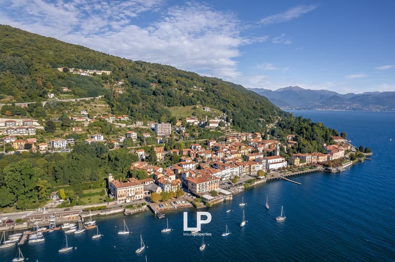 Town surrounded by green hills, waterfront buildings, marina with docked boats, mountains in the distance, clear sky, sea with several boats
