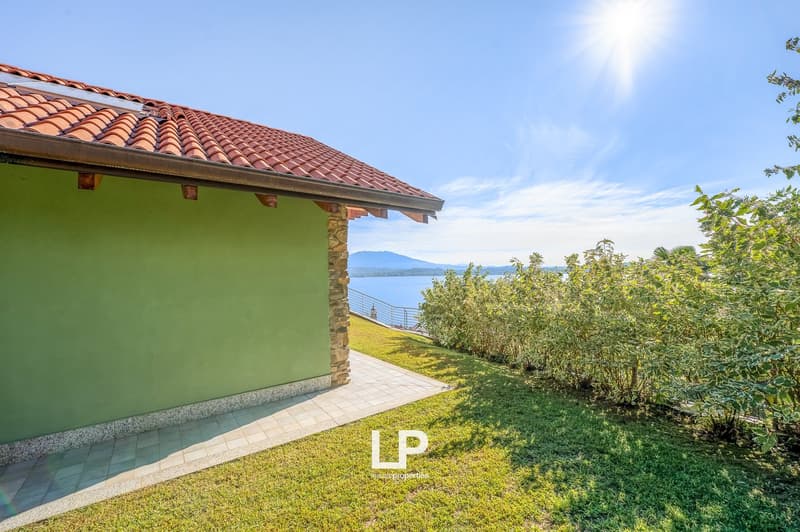 Single story house with green wall and red roof, large balcony with lake view, pathway, and lawn