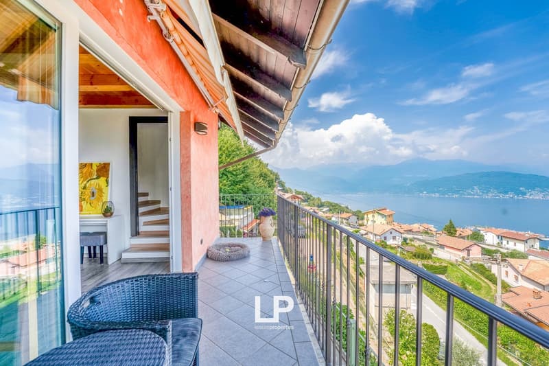 Balcony with black railing, beige tiled floor, two wicker chairs with blue cushions, large windows, mountain and lake view.