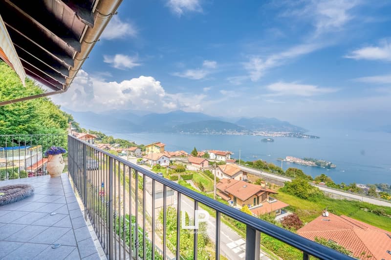 Balcony with tiled floor, metal railings, large view of sea and mountains, vase with flowers, residential area below, clear blue sky with some clouds
