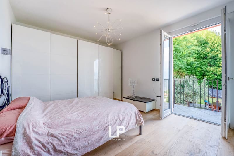 Modern bedroom with white wardrobes, wooden floor, chandelier, double bed with pink bedspread, balcony with fence and view of the outdoors.