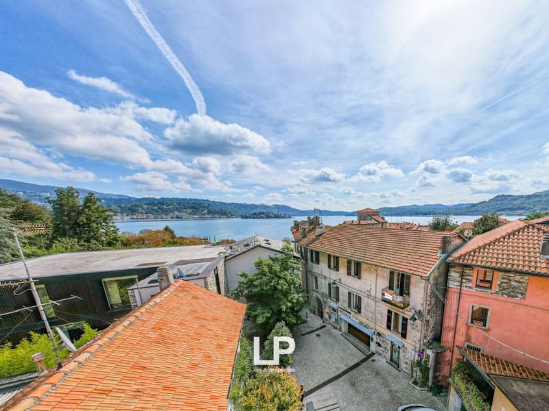 Aerial view of buildings, terracotta roofs, waterfront, mountains in background, clear sky, lake, small trees and shrubs