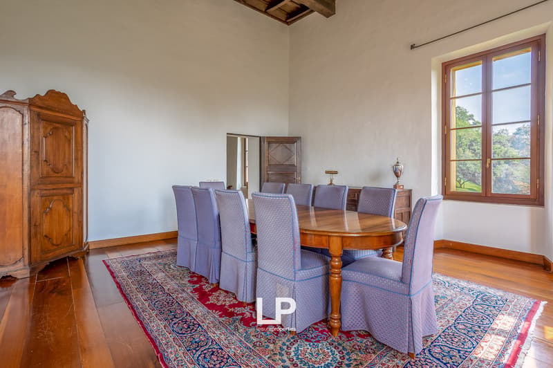 Large wooden dining table, covered with a carpet, eight matching chairs, wooden sideboard, decorative lamps, and a wooden window with a view outside