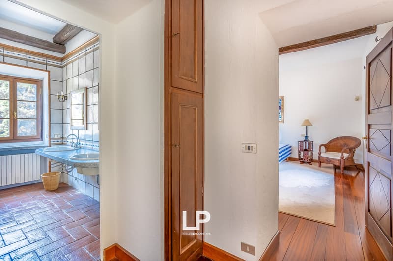 The bathroom features two washbasins, two sinks, and tiled walls. There is a wooden chair and a desk with a lamp. An open doorway leads to another room with a white rug and a bed. The walls are white and there is a power outlet in the wall.