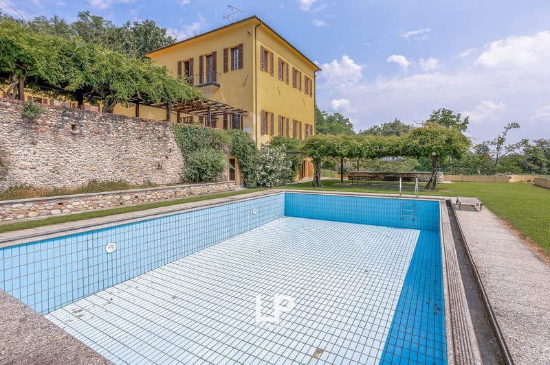 Swimming pool, yellow villa with a garden, stone wall, balcony, trees, green grass, wooden bench, clear sky