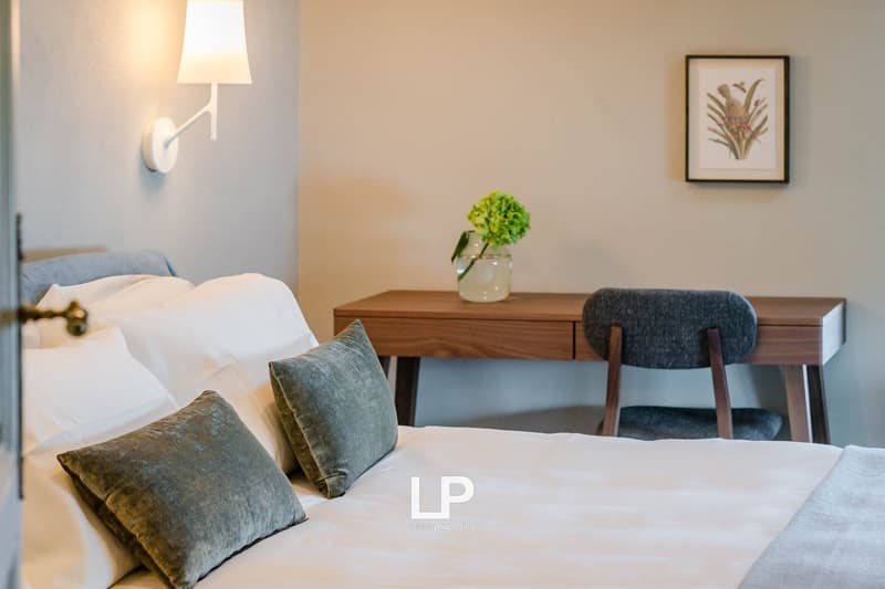 A bed with white sheets and green pillows, a desk with a flower vase, and a chair, a wall lamp, and a framed picture on the wall