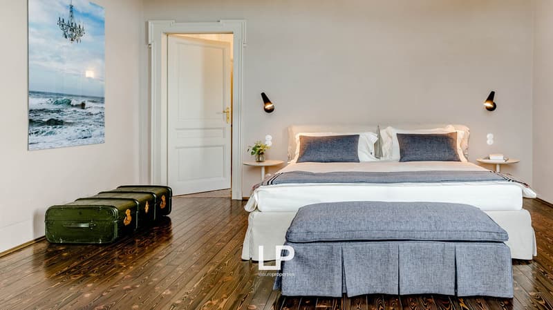 bedroom with white bed, wooden floor, wall lamp, wooden nightstand, ottoman, trunk, painting, door, and small flowers
