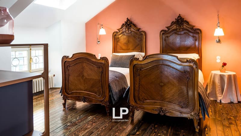 Wooden floors, two beds with ornate wooden headboards, lamps mounted on the wall, white walls, white and pink-orange walls, wooden nightstand, and a radiator.