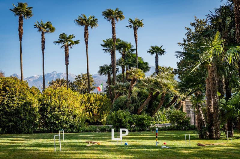 Well-manicured garden with lush green grass, large palm trees, Italian flag, mountains in the background, croquet set-up