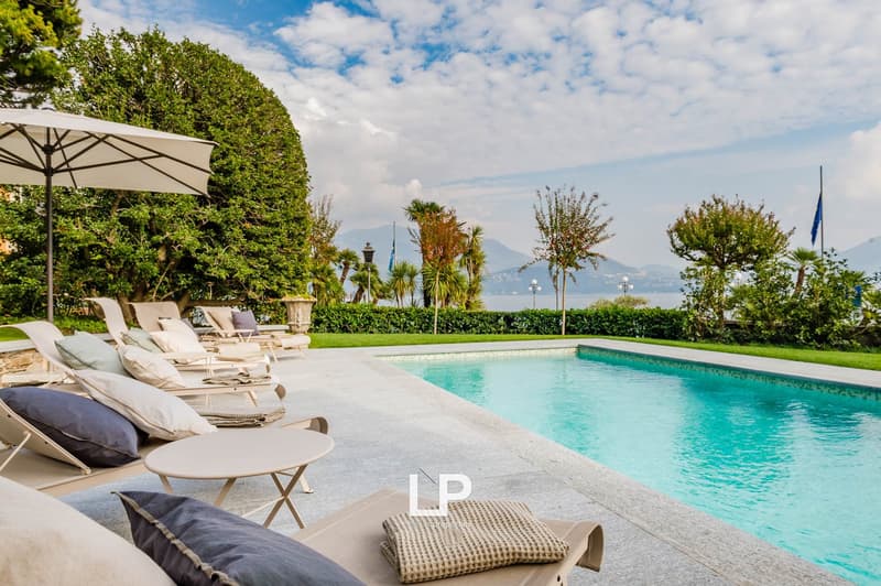 Swimming pool with lounge chairs, umbrella, and table. Green grass, shrubs, and palm trees. View of the sea and mountains.