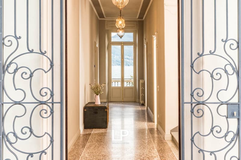 Spacious hallway with double doors, ornate iron gates, wooden chest with flowers, decorative lighting, and view of the lake and mountains.