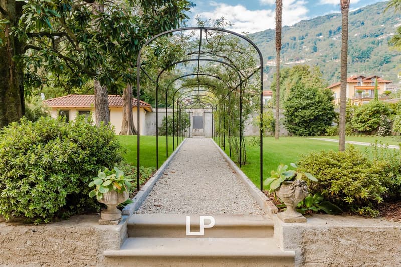 Garden with stone pathway leading to a house, surrounded by bushes, greenery, and mountains in the background.