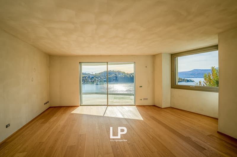 Empty room with wooden floors, sliding glass doors to a terrace, large windows overlooking a lake and mountains, plain walls, ceiling with light reflections.