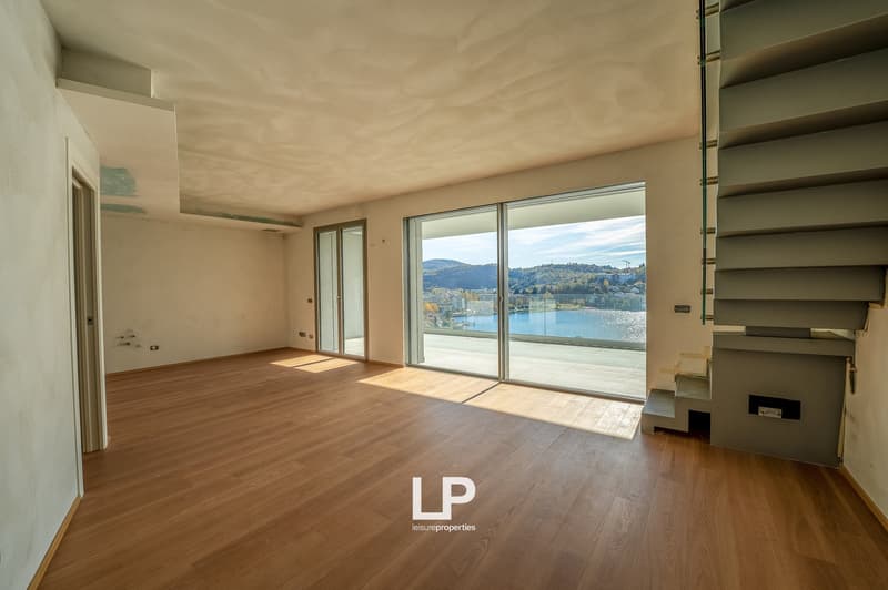 Open room with wooden floor, glass doors leading to balcony with view of lake, mountains, and town
