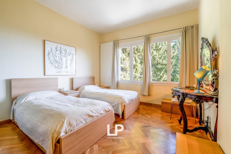 Two beds, nightstands, white bedding, desk with lamp, mirror, windows with curtains, painting