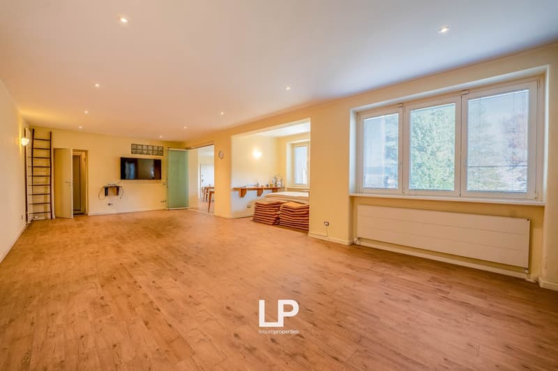 Empty room with hardwood floors, large windows, TV mounted on the wall, door, ladder, heater, large window sill, bright ceiling lights.