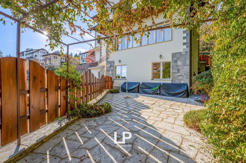 stone paved pathway, wooden fence, small covered patio with loungers, covered with tarps, greenery, building in the background, lampposts, sky, distant buildings