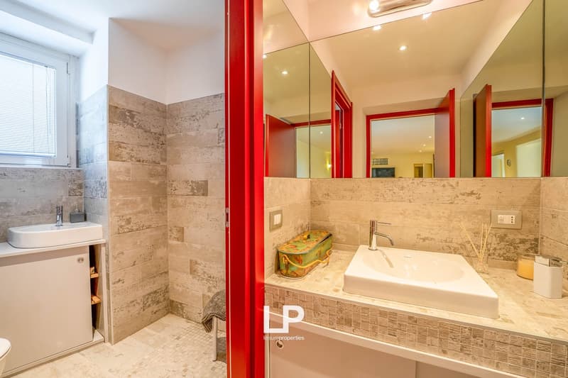 modern bathroom with large mirror, white sinks, grey tiled walls and floor, built-in cabinet, small towel rack, window with blinds