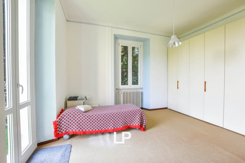 Single bed with red and white bedding, lamp on the ceiling, a nightstand, a small rug, built-in wardrobes, double window, radiator.