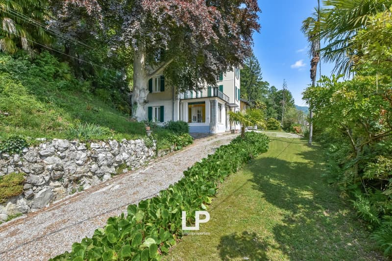 Two-story villa, white exterior, green shutters, stone driveway, lush green landscape, surrounding trees and plants.