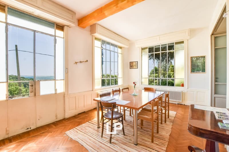 A large dining room with a wooden table and chairs, a rug on the floor, and large windows with a view of trees and the sea in the distance.