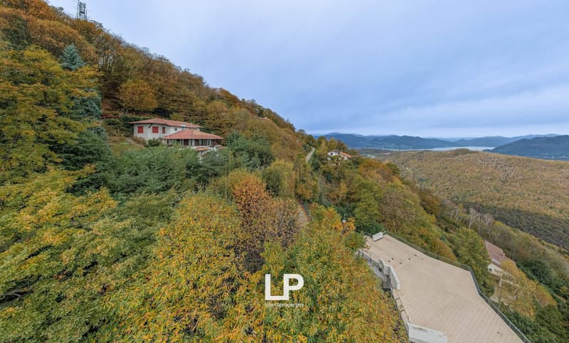 House perched on a hill, white with red roof, surrounded by autumnal foliage, deck, distant mountains, body of water, clear skies